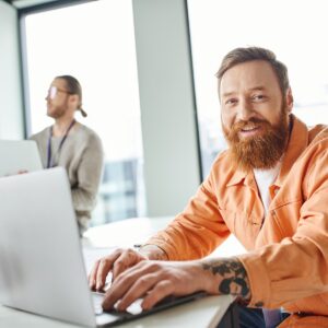 cheerful bearded and tattooed architect working on laptop near takeaway drink in paper cup and looking at camera while colleagues talking on blurred background in coworking design studio